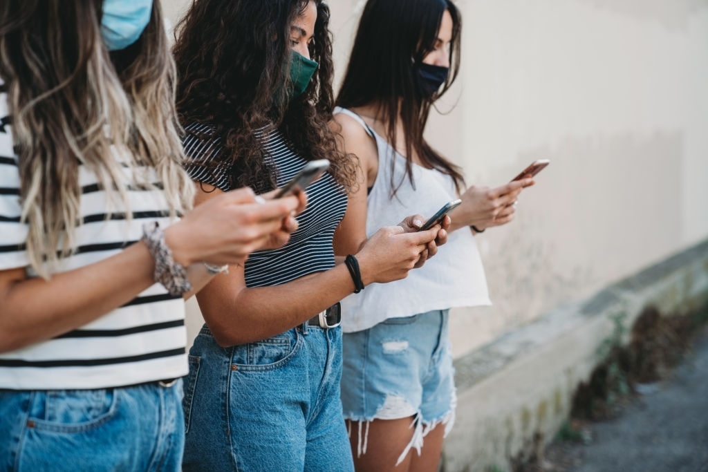 Group of young adult friends standing against a wall, using smart phones and wearing protective face masks. Mixed race and caucasian ethnicities.